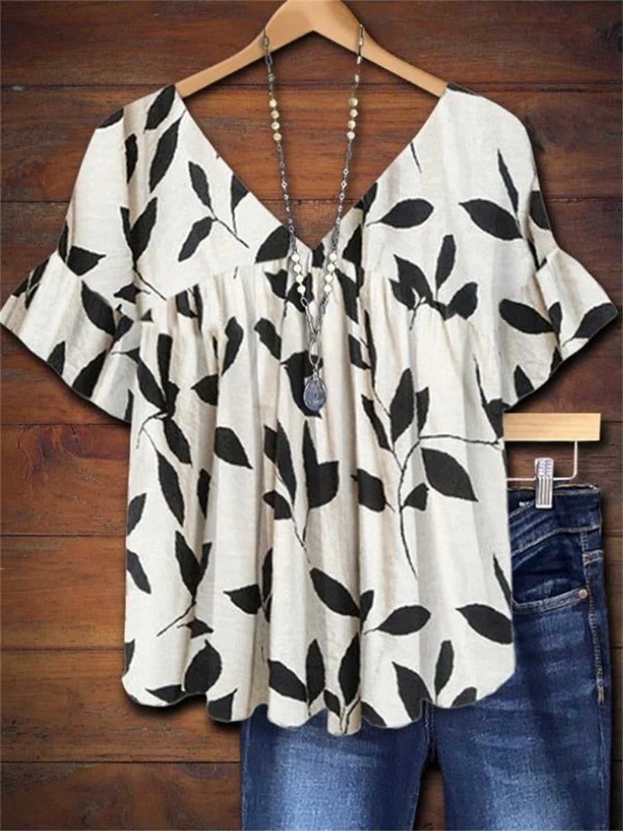 White women's blouse with black botanical leaf print, gathered V-neckline, and short flared ruffle sleeves, shown on a model